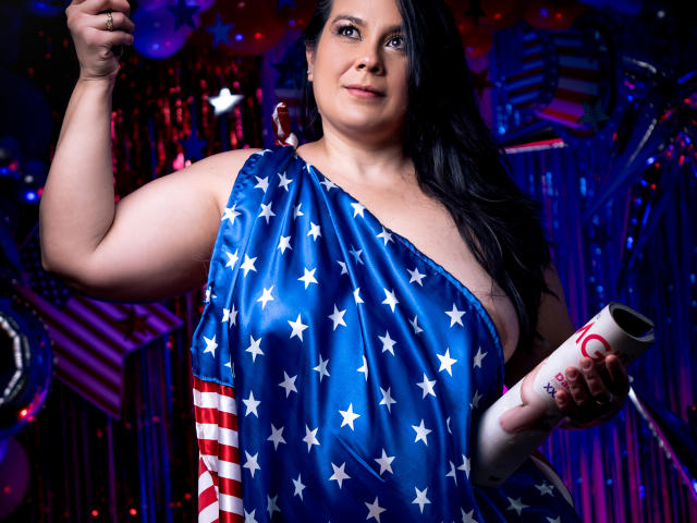 MartinaFox69 with large tits, wearing a blue American flag dress, black hair, holding a scroll, raising her right arm, smiling, in a brightly lit, colorful, and festive setting.