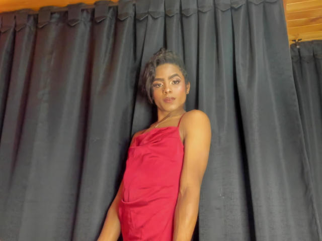 Alaia Clark, in piedi, indossa un abito rosso, ha un braccio muscoloso, capelli ricci neri e un'espressione neutra, davanti a tende scure e a un soffitto in legno..