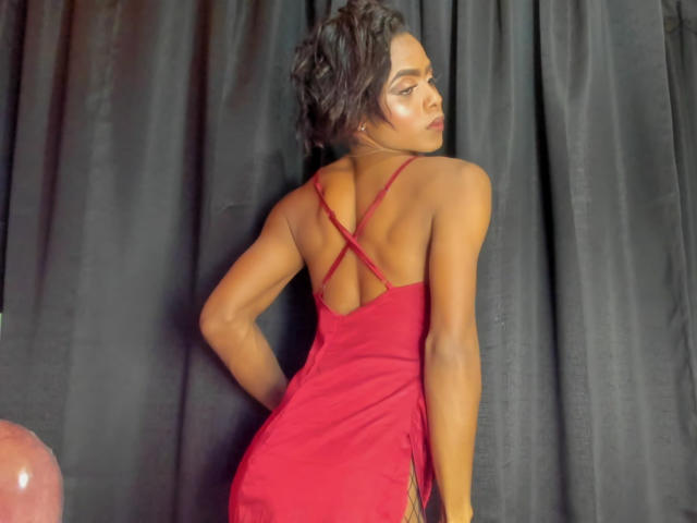 Alaia Clark in un abito rosso, con la schiena rivolta verso la fotocamera, mostra il sedere con spalline incrociate, capelli ricci scuri, pelle luminosa, guarda oltre la spalla, in piedi di fronte a tende nere..