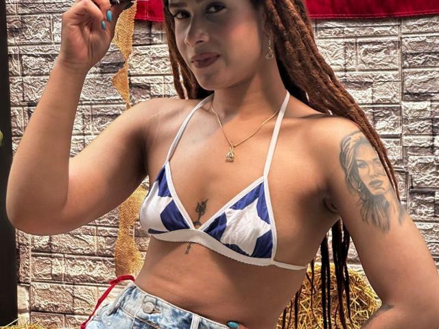 KendallQuinn with brown braids, wearing a blue and white star-patterned bikini top, showing tattoos on her arm and chest, holding a cigarette, and wearing denim shorts.