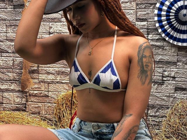 KendallQuinn with brown dreadlocks, wearing a blue and white star-patterned bikini top, denim shorts, and a cowboy hat, showing tattoos on her right arm and chest.