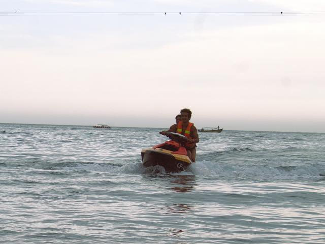 LailaAsher rides a red and yellow jet ski in the ocean, wearing a yellow life vest. Water splashes around her, with a small boat in the distance.