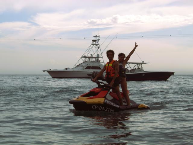 LailaAsher mit kleinen Brüsten und einem schlanken Hintern auf einem roten und gelben Jetski, trägt eine orangefarbene Schwimmweste, während eine andere LailaAsher in einem Boot die Hand hebt..