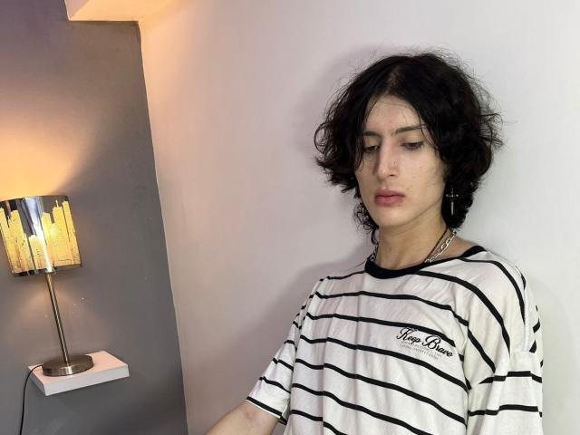 Asmodues met rommelig zwart haar, die een wit gestreept shirt, zwarte oordopjes en een ketting draagt, staat in de buurt van een brandende lamp en kijkt naar beneden met een neutrale uitdrukking..