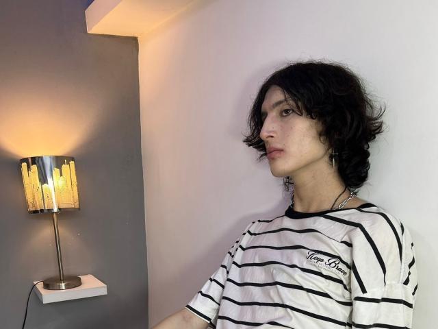 Asmodues with messy black hair, wearing a white striped shirt, standing near a modern lamp, looking to the side with a neutral expression, soft light on their face.