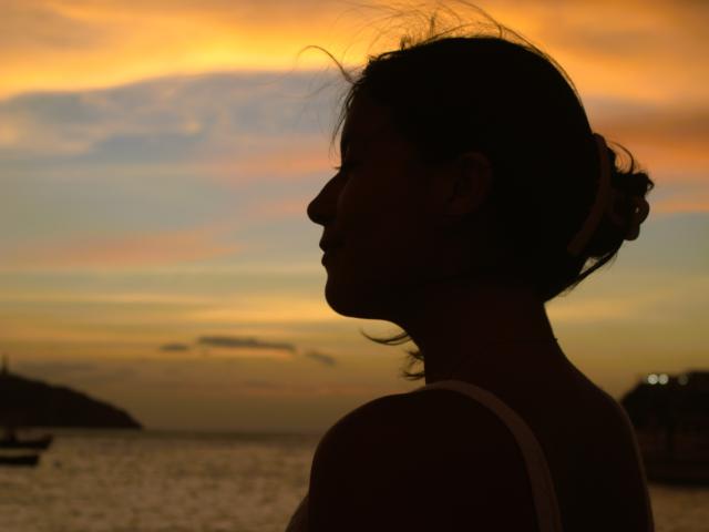 LailaAsher, con i capelli scuri raccolti in un disordinato chignon, indossa una canotta bianca e guarda a destra, sullo sfondo di un tramonto colorato. Il suo profilo mostra un seno leggermente pronunciato e un'espressione serena..