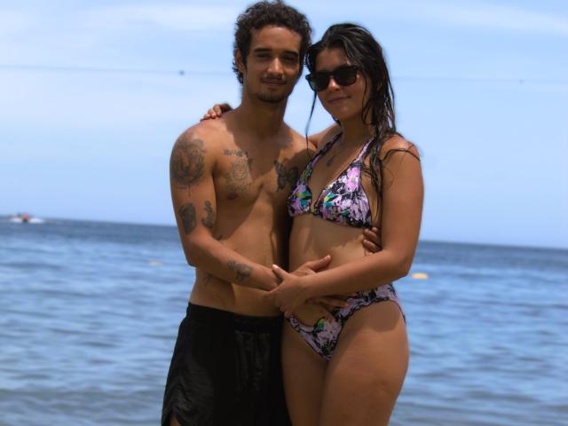 LailaAsher, avec ses cheveux bruns bouclés, ses tatouages et son short de bain noir, se tient à côté de LailaAsher, qui porte un bikini fleuri, et elles se tiennent la main, toutes les deux portant des lunettes de soleil, au bord de l'océan bleu..