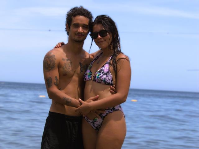 Laila Asher y su pareja, ambos sin camisa, ella con los senos cubiertos por un bikini floral rosa y negro, tomados de la mano en una playa con un océano azul de fondo..