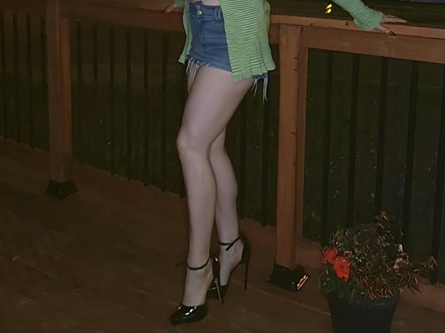 TitanicTits, avec des jambes chaussées de talons hauts, portant un cardigan vert rayé et un short en jean, se tient sur un balcon en bois avec une plante en pot et une rambarde sombre..