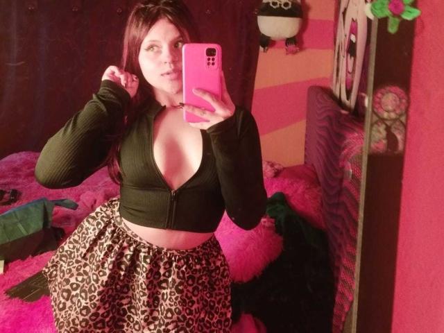 SophieHill with red hair, black zip-up top showing her tits, and leopard print skirt, taking a mirror selfie with a pink phone, holding her hair.