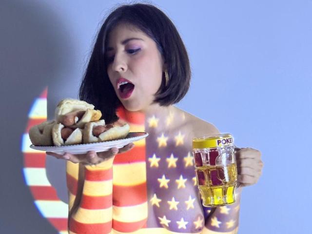 AnastasiaEvansX with star and stripe body paint, holding a beer and hot dog, mouth open, looking surprised. She's got a short black bob and red lipstick.