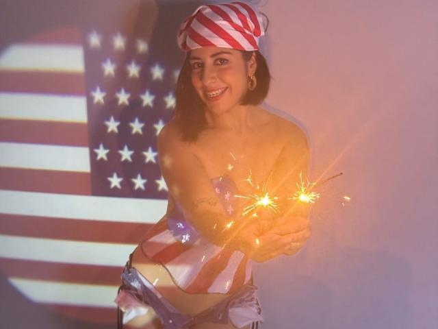 AnastasiaEvansX with red and white striped hat, holding sparklers, smiling, wearing a star-spangled top, and silver shorts, showing her tits, with an American flag projection behind her.
