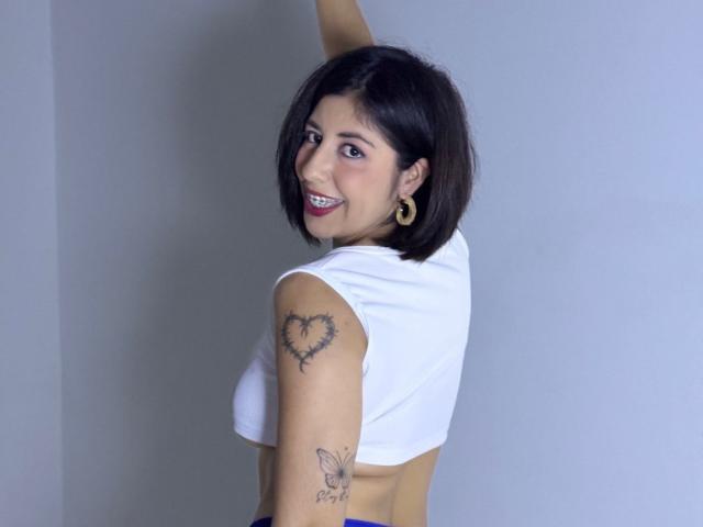 AnastasiaEvansX with short black hair, smiling, wearing a white crop top, showing a heart tattoo on her right arm and a butterfly tattoo on her left. Boobs visible.
