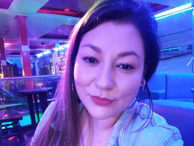 SakotoDika, con lunghi capelli castani, rossetto rosso e un trucco leggero, indossa una camicia di jeans, sorride in un bar illuminato al neon, mostrando il suo viso carino e il suo sedere piccolo e rotondo..