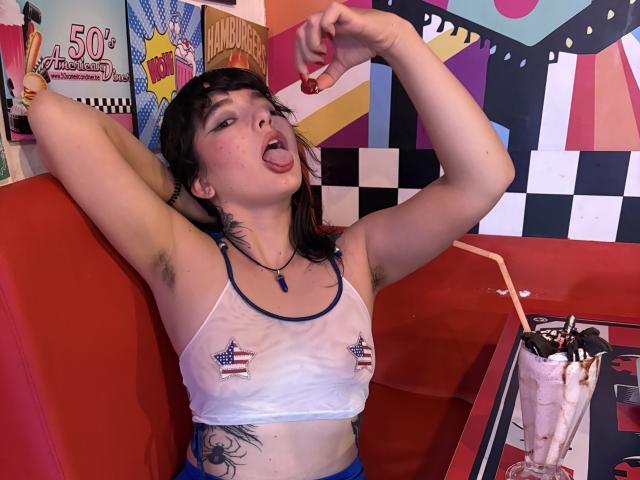 HairyValeri, with arms raised, tongue out, wearing a white crop top with American flag nipples, in a red booth, has a tattoo and a milkshake with a straw.