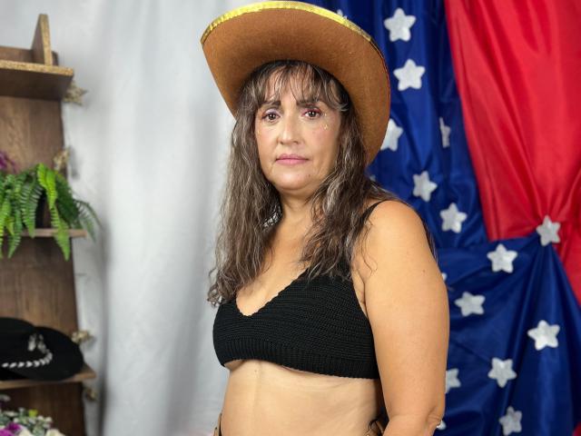 SashaStonne with wavy brown hair, tan skin, black bra, showing small tits, wearing a brown cowboy hat, standing in front of a blue and red flag.