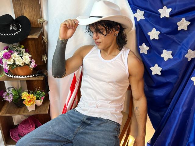 AlexBernal mit zerzaustem, braunem Haar, weißem Cowboyhut, Tattoostreifen am Arm, weißem Tanktop, blauen Jeans, sitzend, rechter Arm erhoben, amerikanische Flagge, Holzregal mit Blumen und schwarzem Hut..