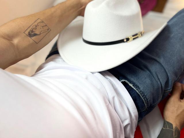 El brazo de AlexBernal tiene un pequeño tatuaje cuadrado. Lleva un sombrero blanco, una camisa blanca y pantalones vaqueros azules. Las manos están apoyadas en la parte baja del abdomen de AlexBernal, mostrando un tatuaje en la muñeca de AlexBernal..