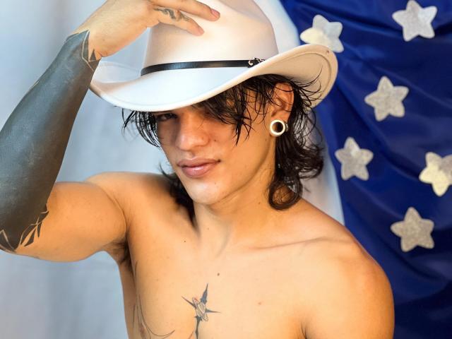 Shirtless AlexBernal with tattoos, white cowboy hat, black arm tattoo, black hair, ear gauges, and a star tattoo on chest, holding hat, wearing a slight smirk, blue and white flag.