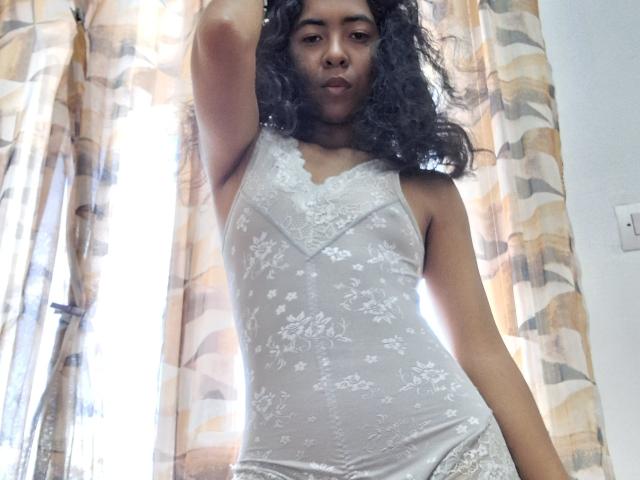 Rosiia, com cabelos escuros e cacheados, vestindo um vestido branco de renda transparente, fazendo beicinho com o braço direito levantado, mostrando seus pequenos seios e nádegas planas. A luz filtra através de cortinas estampadas..