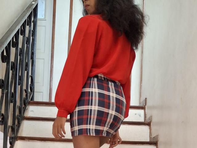 Rosiia with curly black hair, wearing a red long-sleeve shirt and a blue, red, white plaid skirt, standing on white stairs with a black iron railing.