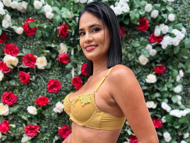 LaylaRoa, con un seno di media grandezza, indossa un reggiseno di pizzo giallo, sorride con rossetto rosso, ha i capelli neri e gli occhi marroni, ed è in piedi di fronte a un muro di rose rosse e bianche..