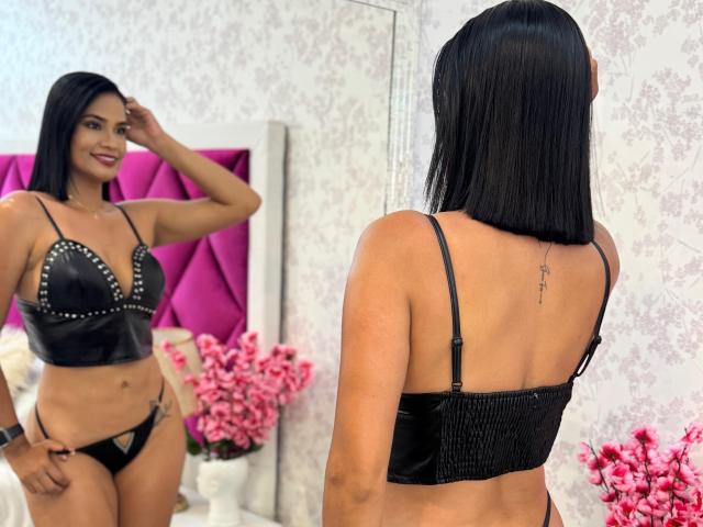LaylaRoa, portant un soutien-gorge et une culotte en cuir noir, montre son dos avec un tatouage, face à un miroir, souriante, debout devant une tête de lit rose, avec des fleurs roses..