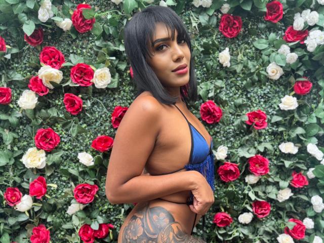 LaylaJones with dark bob, wearing a blue bikini top, showing a large tattoo on her right hip, standing in front of red and white roses. Her tits are small.