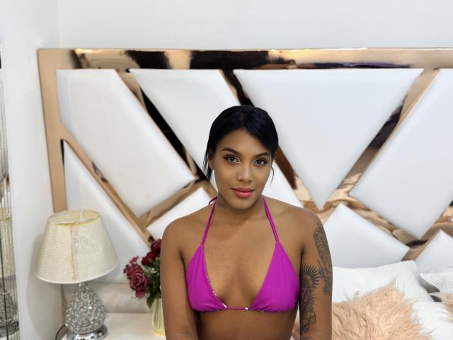 Layla Jones, con piel de tono marrón medio, cabello negro y un top de bikini rosa que muestra pequeños senos y un tatuaje en su brazo derecho, está sentada en una cama con un cabecero metálico..