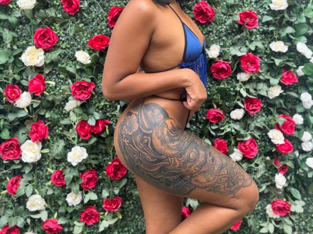 Layla Jones, usando um biquíni azul, exibindo uma bunda grande com tatuagens intrincadas em tinta preta, em frente a rosas vermelhas e brancas..