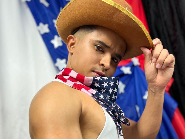 DayanWilly, com a pele bronzeada, usando um chapéu de cowboy e um cachecol com a bandeira americana, segurando o chapéu, mostrando um braço musculoso e uma expressão séria. Seus olhos castanhos estão fixos para frente..