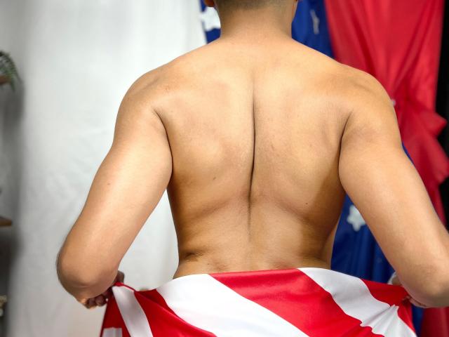 La espalda desnuda de DayanWilly, con la columna vertebral visible, sosteniendo una bandera roja y blanca, brazos musculosos, cabello corto y oscuro, piel bronceada, de pie con la bandera cubriendo sus glúteos..