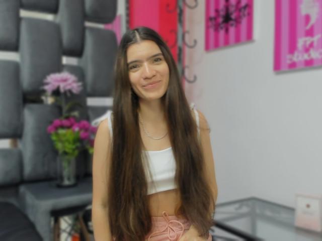 SaraOwenn, con cabello castaño largo, pechos pequeños, vestida con un top corto blanco y una falda rosa con cordones, sonriendo en una habitación con paredes acolchadas negras y decoraciones rosas..