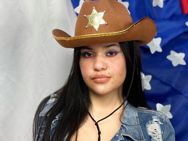 CloverLover with long black hair, brown cowboy hat, and gold sheriff star, wearing a blue ripped denim jacket, and a black necklace against a white and blue background.