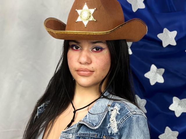 CloverLover with long black hair, wearing a brown cowboy hat with a sheriff's badge, and a ripped denim jacket, has pink eyeliner and a small beauty mark on her cheek.