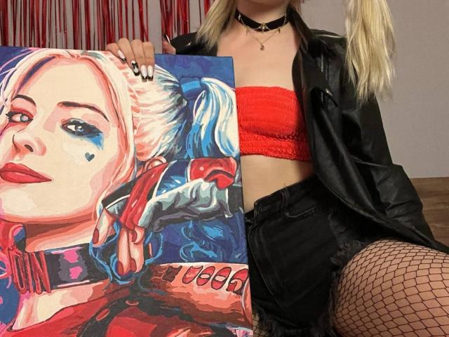 LaPulia mit blonden Zöpfen, die ein rotes Top, eine schwarze Lederjacke und Fischernetzstrümpfe trägt, hält ein farbenfrohes Gemälde von sich selbst, auf dem sie blaues Augen-Make-up und ein Herz zu sehen ist..
