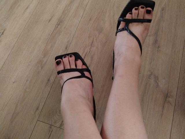 DeesseHelena's feet in black strappy high heels with dark red toenails, standing on wooden floor. Her ankles are slightly crossed, showcasing the sleek design of the sandals.