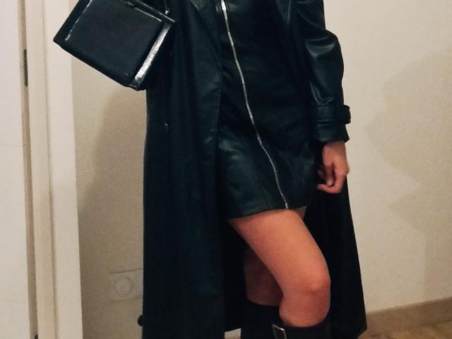 DeesseHelena wearing a black leather dress with a zipper, black boots, and a black leather purse, showing her thighs and butt. Her legs are slightly apart.