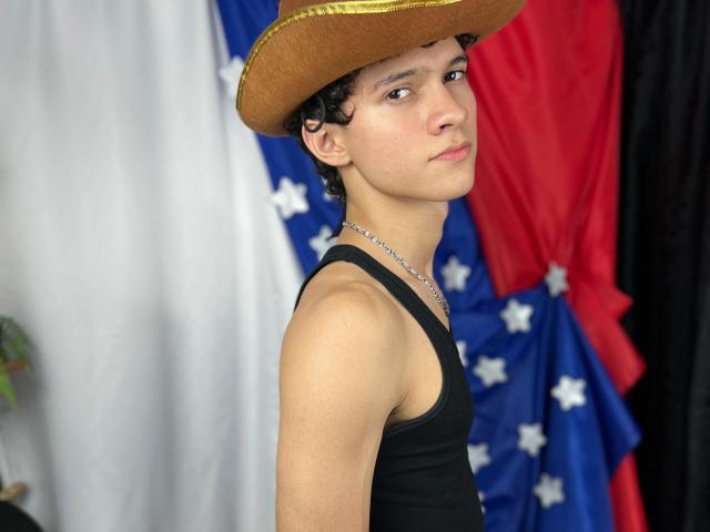 SamuelHills with curly black hair, brown cowboy hat, black tank top, silver necklace, and a serious look, standing in front of a blue, white, and red flag.