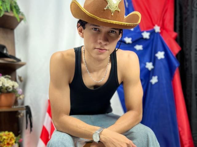 SamuelHills draagt een cowboyhoed met een ster, een zwart tanktop, blauwe jeans, een zilveren ketting en een horloge, en zit met een serieuze uitdrukking, kijkend naar de camera..