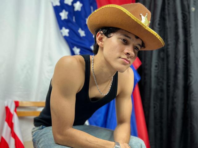 SamuelHills, wearing a brown cowboy hat, black tank top, and blue jeans, leans forward with a pensive expression, showing a muscular, tan, handsome face and a silver necklace.