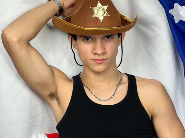 SamuelHills, met een bruine sheriffshoed met een gouden ster, een zwart tanktop en een zilveren ketting, heeft een neutrale uitdrukking, met een arm omhoog, waarbij okselhaar zichtbaar is..