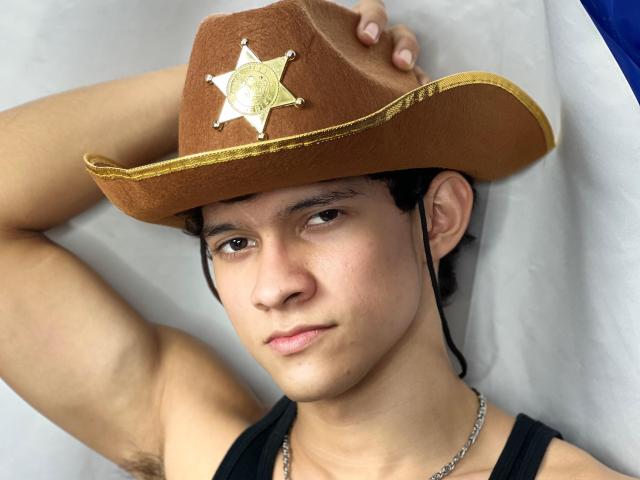 SamuelHills, met een bruine sheriffshoed, bruine ogen, zwart haar en een lichte glimlach, draagt een zwart tanktopje, waar je okselhaar zichtbaar is, en een zilveren ketting..