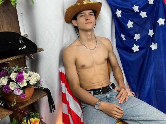 SamuelHills zonder shirt, met een cowboyhoed, gekleed in jeans en een riem, zittend tegen een vlag, met een zwarte hoed en bloemen aan de linkerkant, met een zelfverzekerde blik..