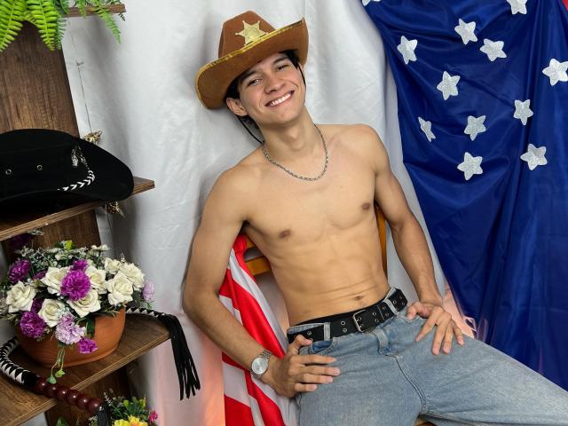 SamuelHills zonder shirt, met een cowboyhoed, lachend, gekleed in jeans en een riem, heeft een slank, gespierd lichaam en draagt een ketting. Op de achtergrond zijn een Amerikaanse vlag en bloemen te zien..