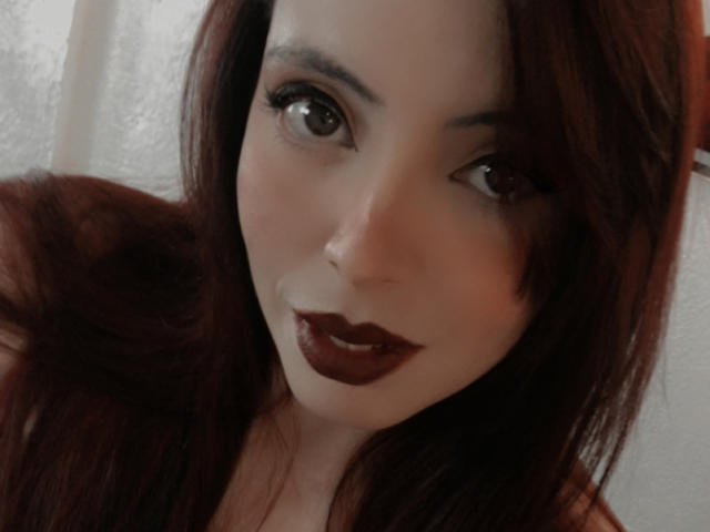 Rosalinnes with dark red hair, brown eyes, and dark lipstick, looking at the camera with a subtle smile, soft light on her fair skin. Her tits are partially visible.