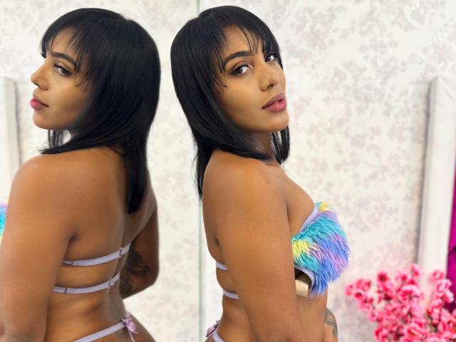 Layla Jones, con piel de tono marrón medio, cabello negro de largo medio, mirando hacia atrás, vestida con un top sin tirantes adornado con plumas de colores, que muestra sus pequeños senos firmes y un tatuaje en su brazo derecho..