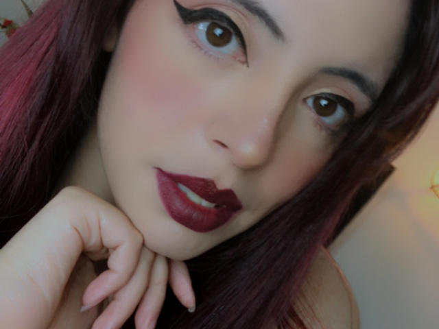 Rosalinnes has red lipstick, dark eyeliner, and pinkish skin. She's looking at the camera with her hand near her chin, and she has pinkish-red hair.