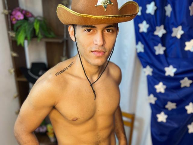 DereckStorm, shirtless, wearing a brown cowboy hat with a metal emblem, black necklace, and a tattoo on his right shoulder, has a muscular build and brown eyes, looking at camera.