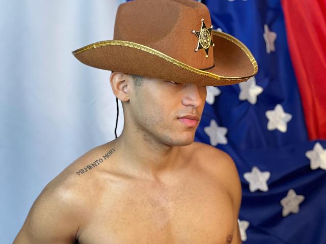 DereckStorm wearing a brown cowboy hat with a sheriff's badge, shirtless, showing a tattoo on the right shoulder, and standing in front of a blue and red flag.